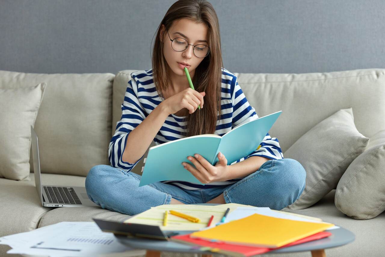 Jovem estuda concentrada conteúdo de livro em sua mão sentada no sofá diante livros e cadernos 