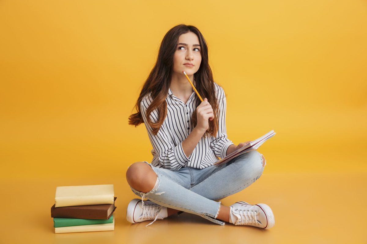 Adolescente sentada no chão aos lados de pilha de livros estudando com um caderno no colo e um lápis na mão encostando no queixo com expressão pensativa 