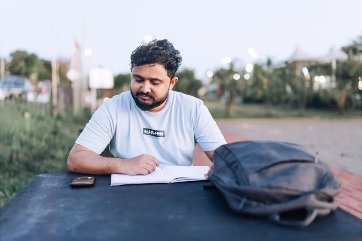 Um estudante em um banco da praça escrevendo no caderno. Em cima da mesa há uma mochila