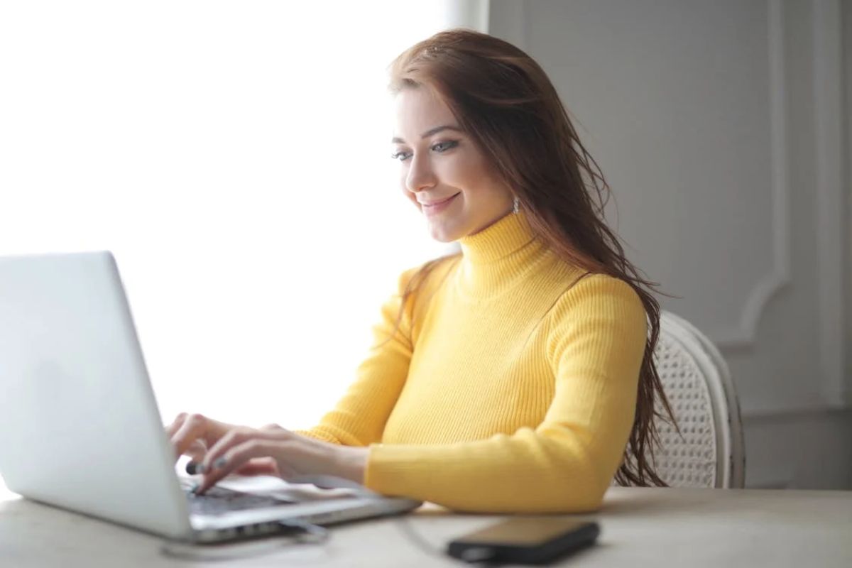 Uma estudante de camisa amarela em casa, ela usa seu notebook apoiada na mesa 