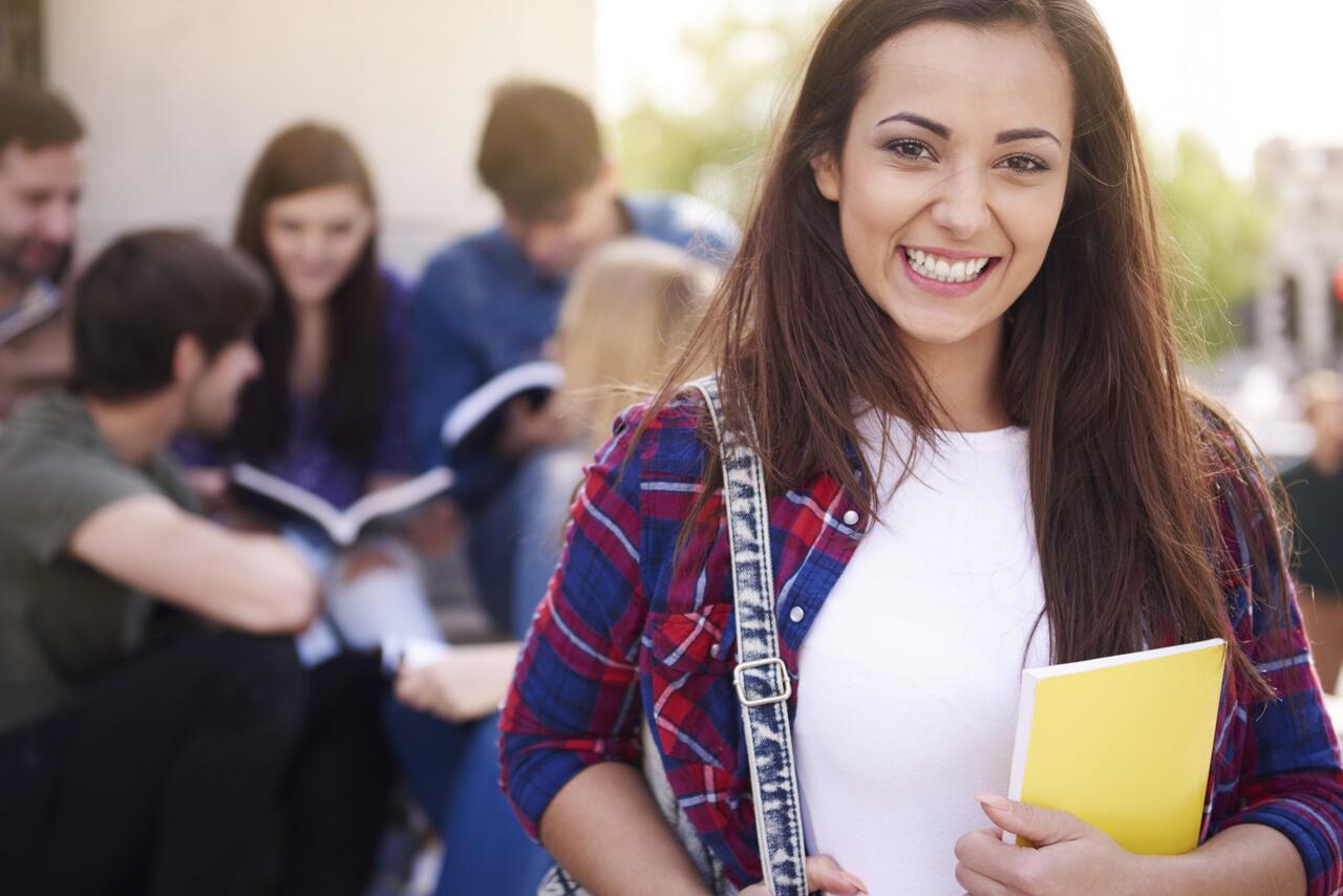 Adolescente sorrindo segurando livros com colegas ao fundo 