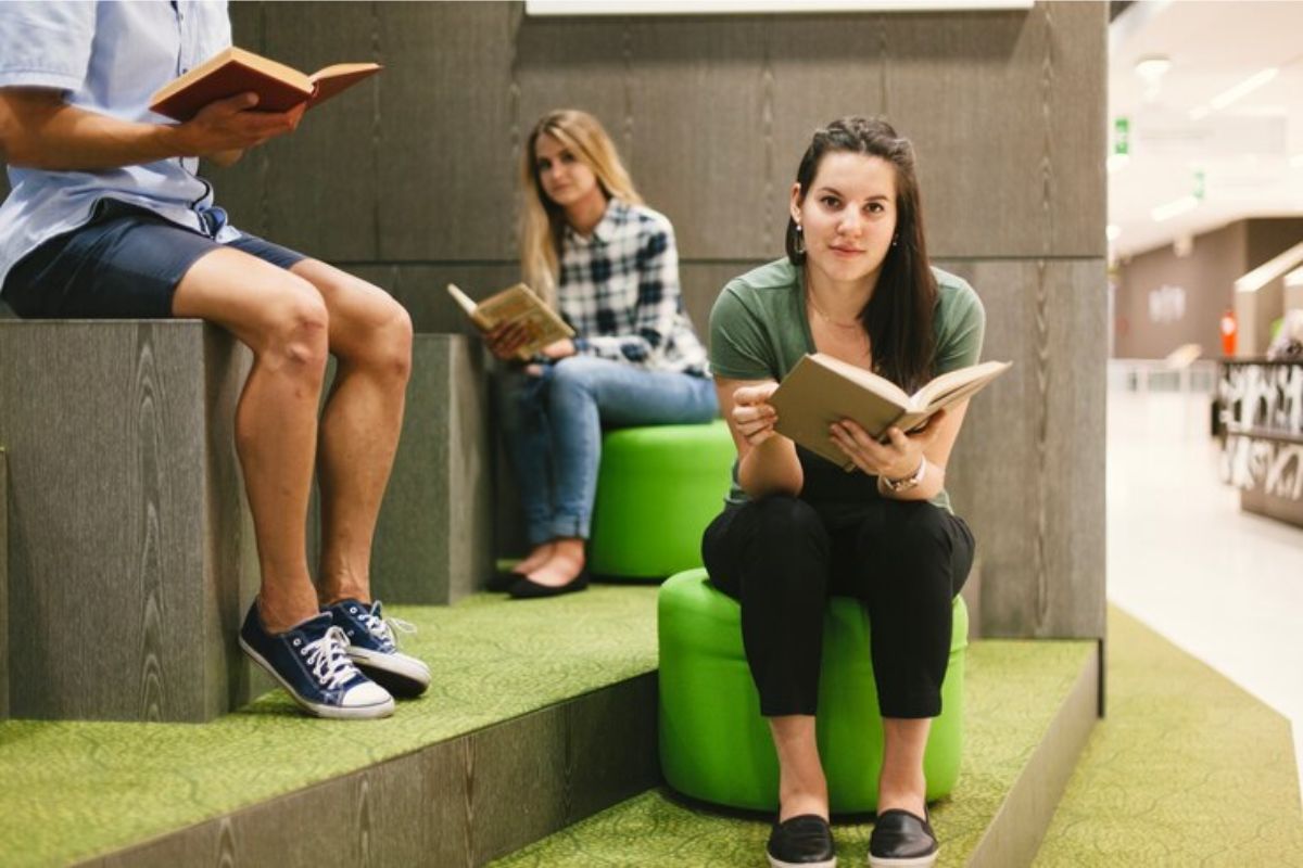 uma estudante com um livro aberto na mão sentada em um puff verde. Ao fundo é possível ver outra aluna lendo também