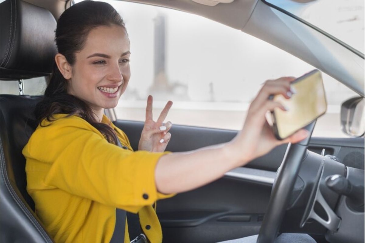 Uma mulher dentro do carro tirando foto com o celular na mão fazendo sinal de V