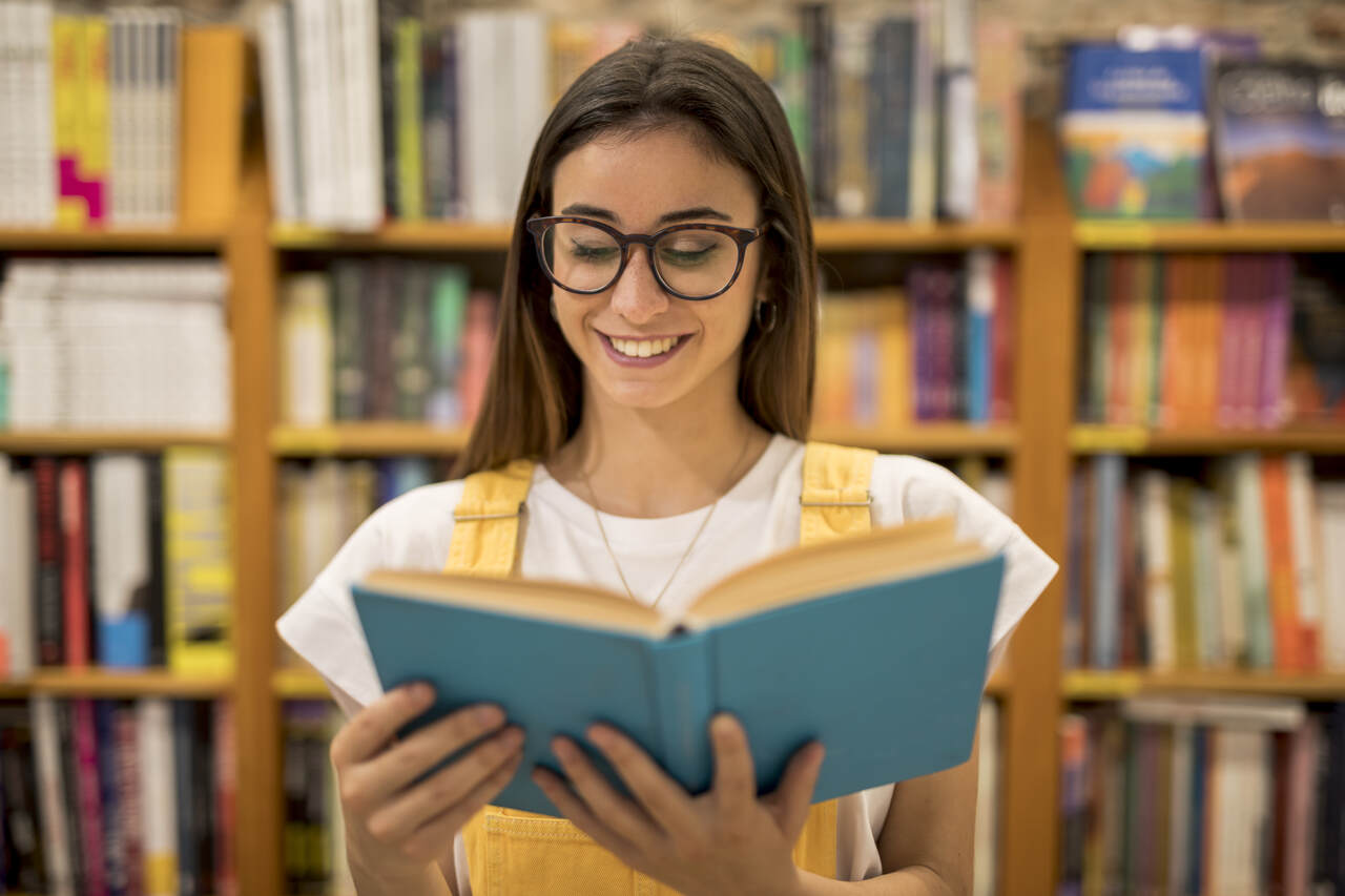 Jovem sorri lendo livro aberto em sua mão em uma biblioteca 